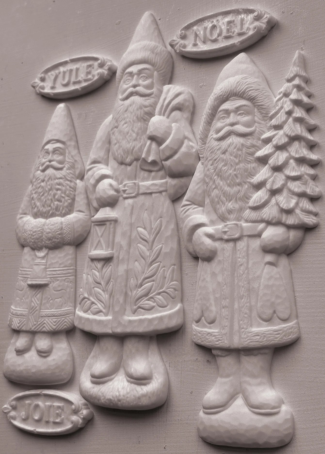 Three carved Santa Claus figures in a relief style, labeled Yule, Noel, and Joy, on a textured gray background.