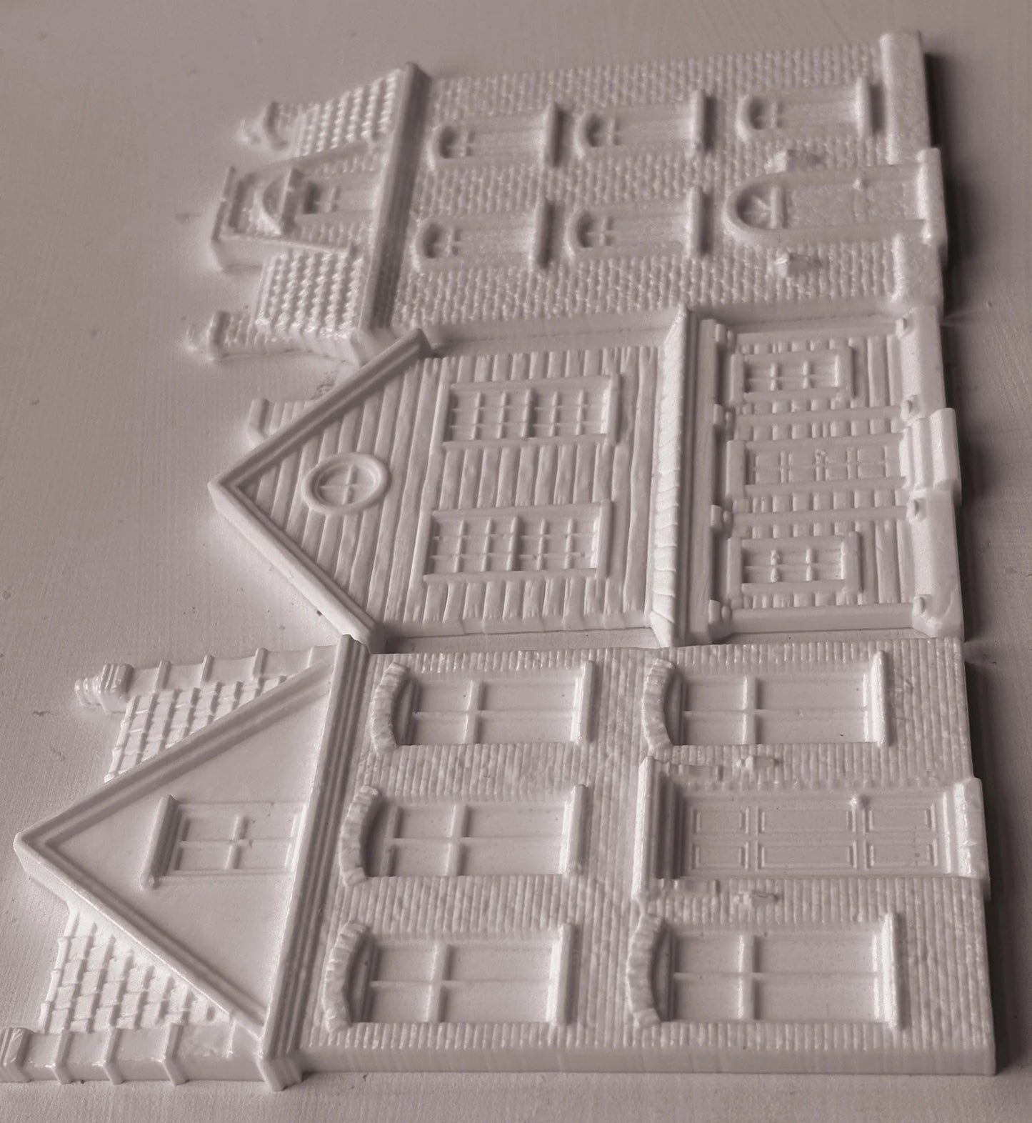 White plastic miniature building pieces for dioramas, featuring detailed house designs with windows, doors, and textured brick patterns, arranged on a light gray background.