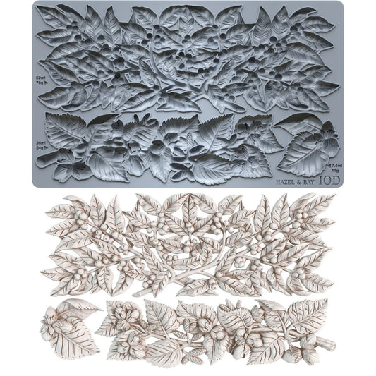 A floral leaf embossing plate made of gray metal, shown above a matching set of embossed leaf and berry cutouts in an off-white tone. Small printed markings on the plate include 'HAZEL & BAY' and '10D'.