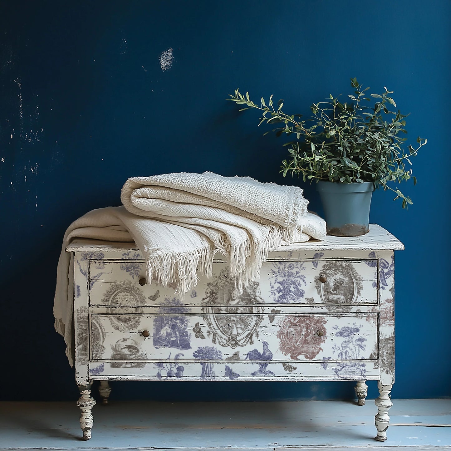 Decorative chest with floral patterns, white blanket, and potted plant against a blue wall.