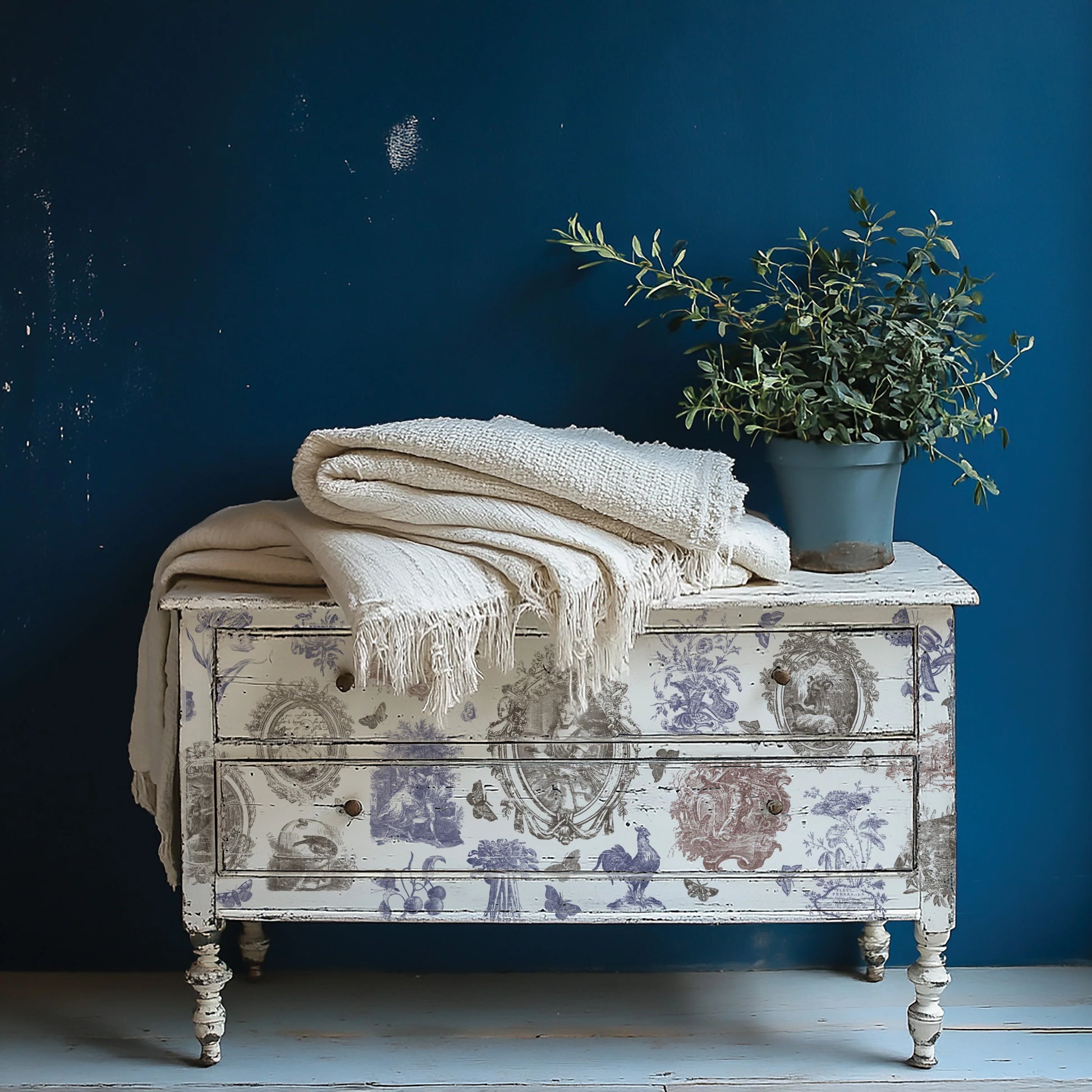 Decorative chest with floral patterns, white blanket, and potted plant against a blue wall.