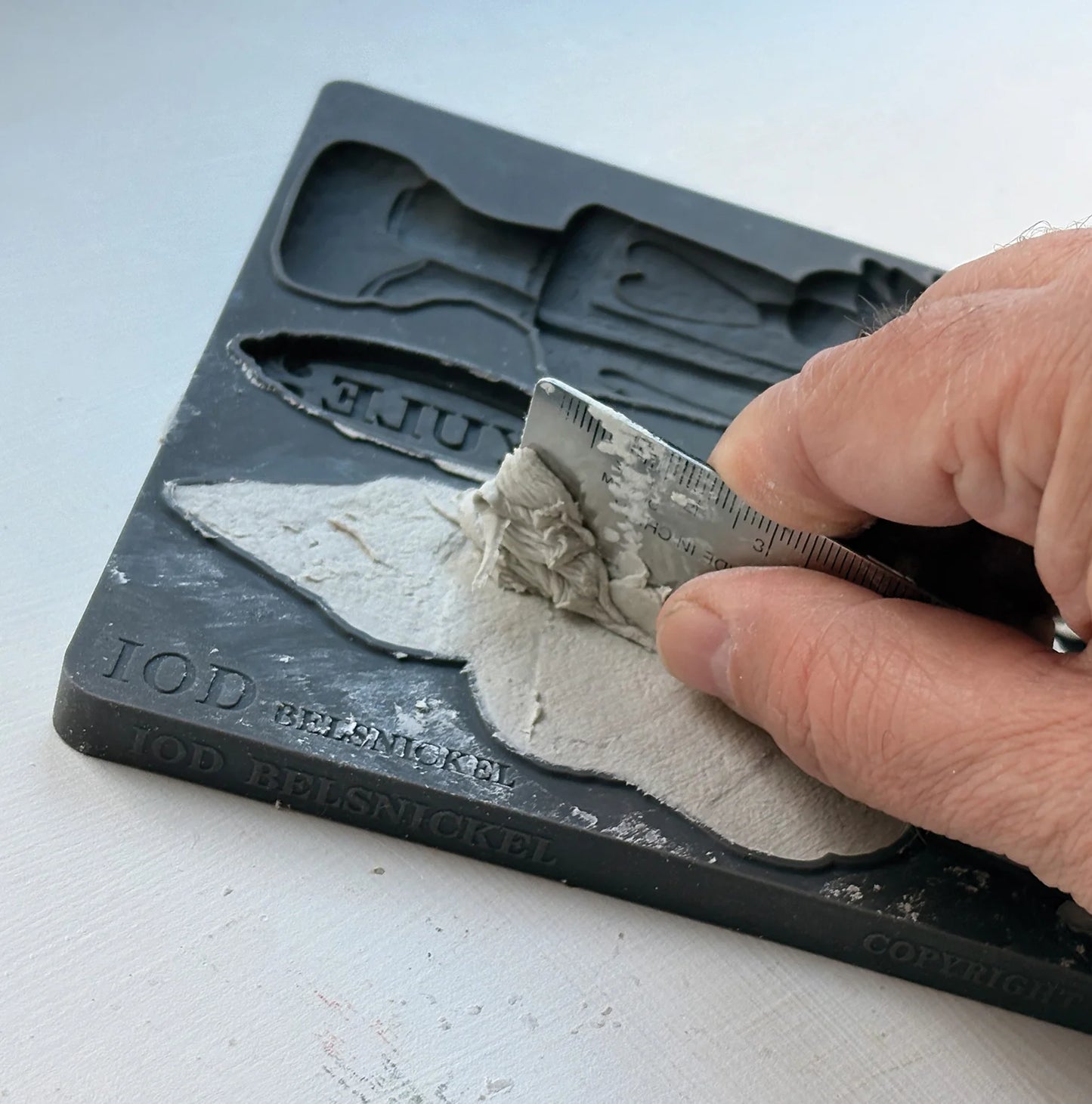 A close-up of a hand using a metal spatula to scrape light gray plaster or clay into a black mold tray, with embossed text including 'TOD Belsmoke' and 'JUEU' visible on the tray.