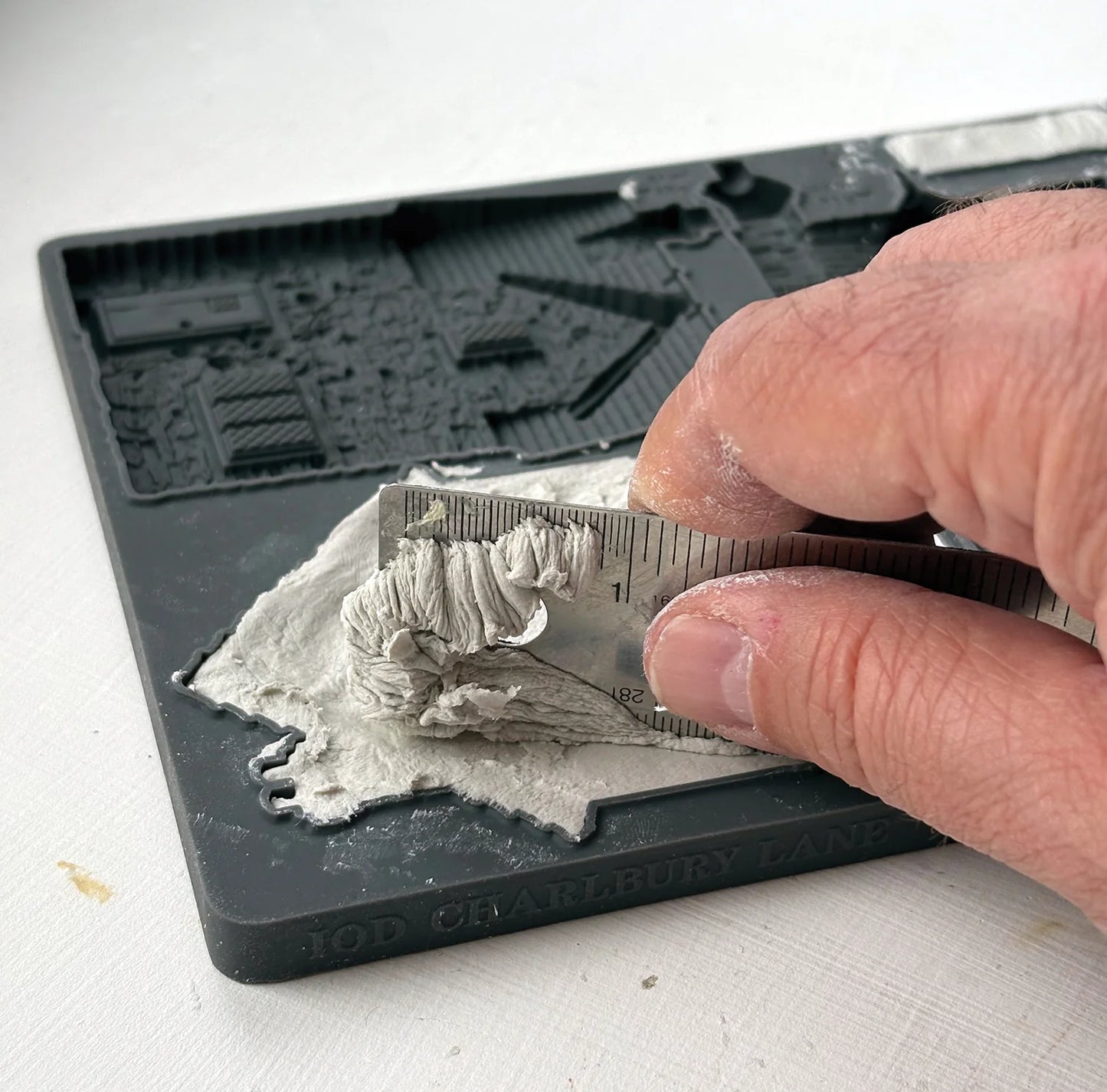A close-up of a hand using a metal ruler to measure a layer of gray plaster or clay on a black 3D printable terrain mold tray.