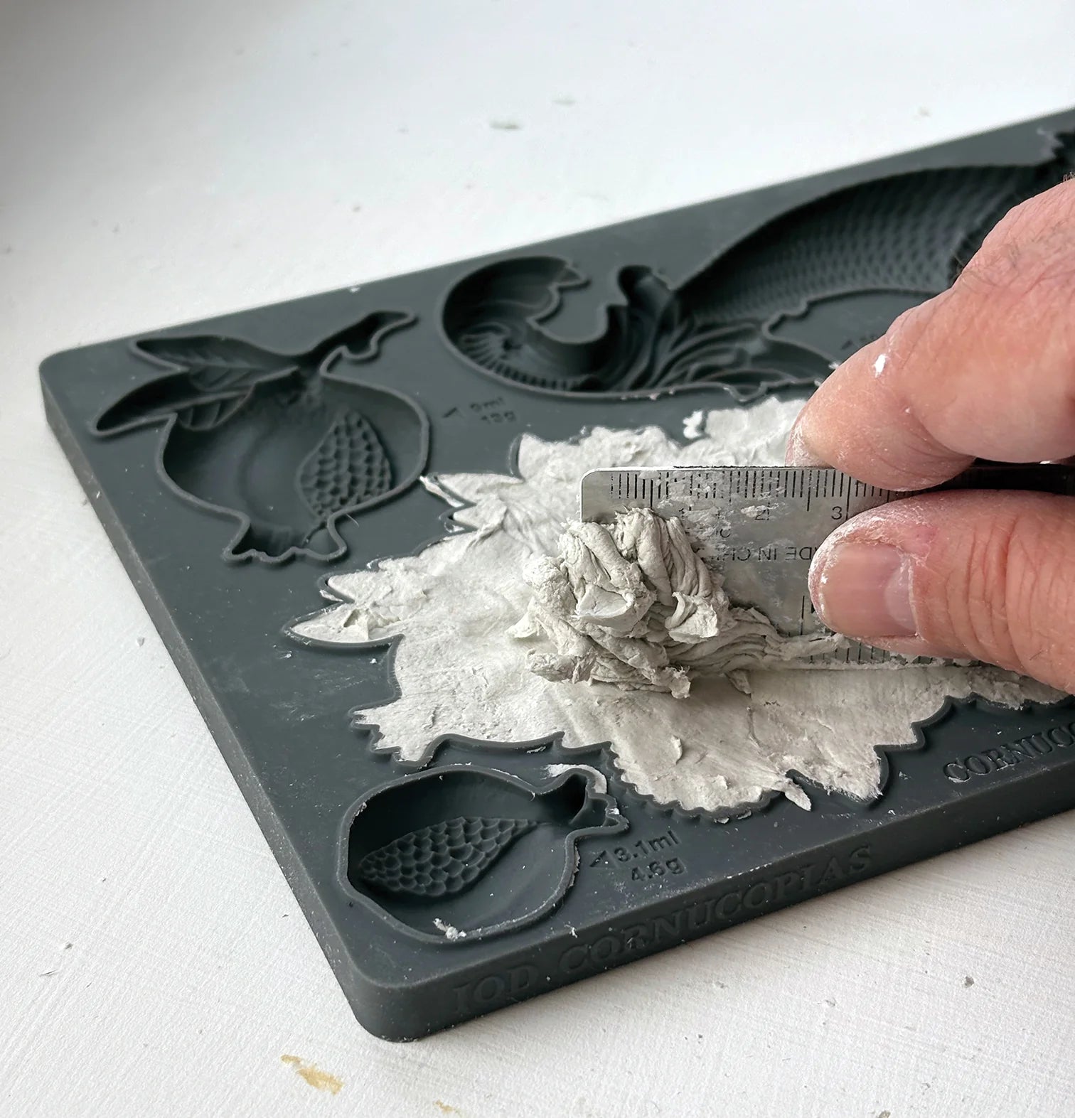 A close-up of a hand using a small metal scraper to remove wet gray plaster or clay from a dark gray silicone mold tray with fruit-shaped cavities.