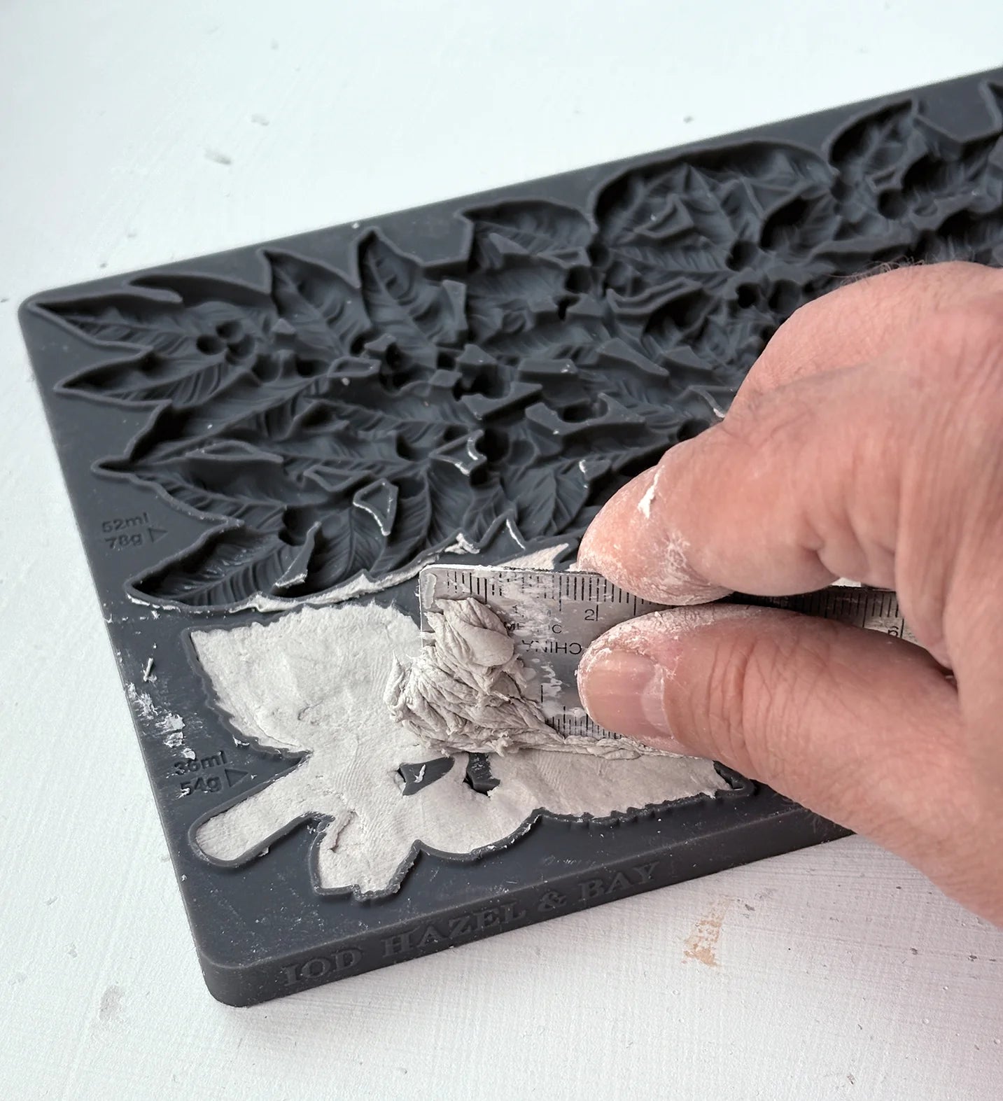 A close-up of a hand applying wet clay to a dark gray plastic leaf mold, showing the mold's floral and leaf patterns.
