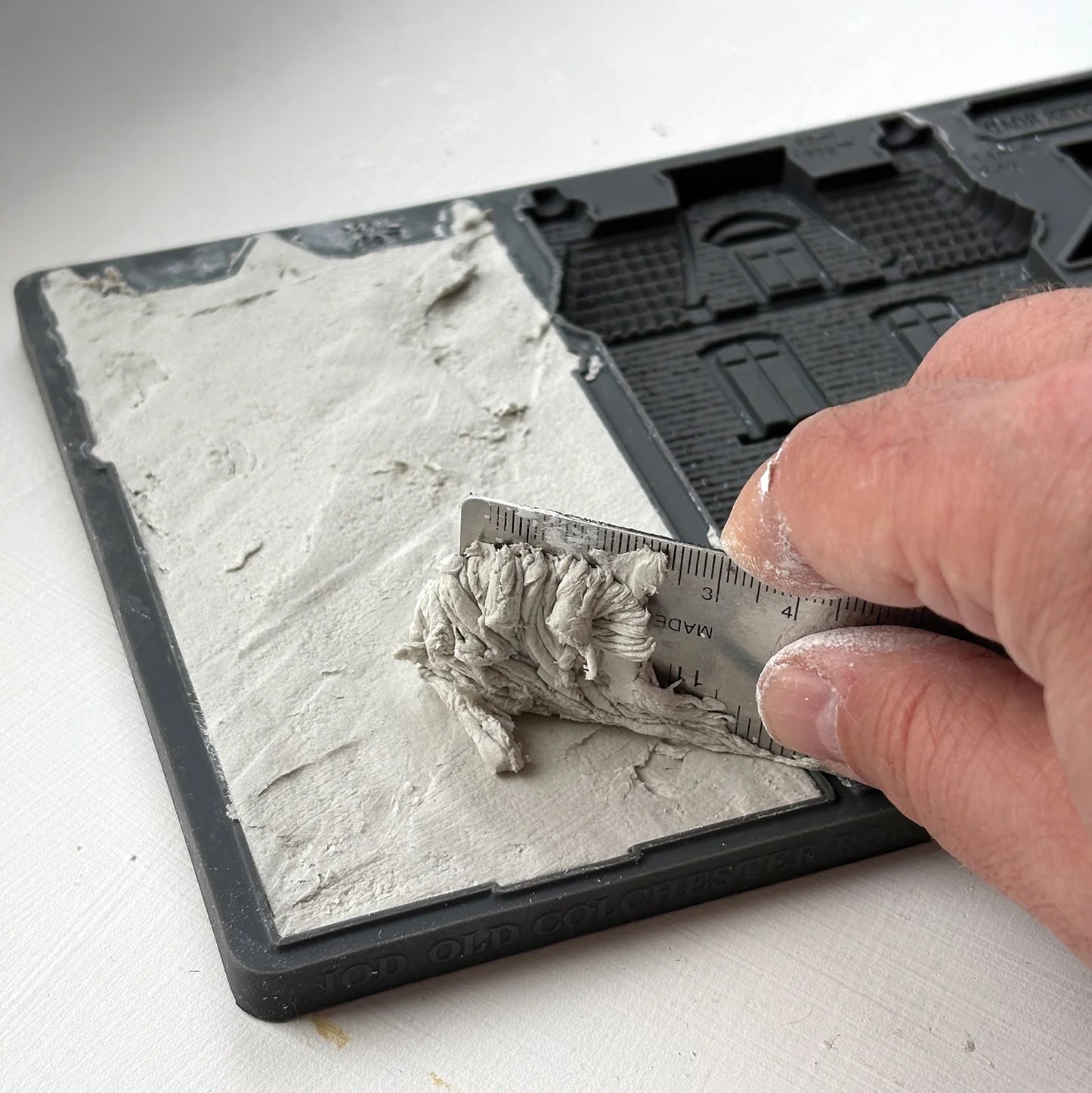 A close-up of a hand using a metal ruler to measure a layer of gray plaster or clay on a black square tray, with the textured material spread across one half of the tray.