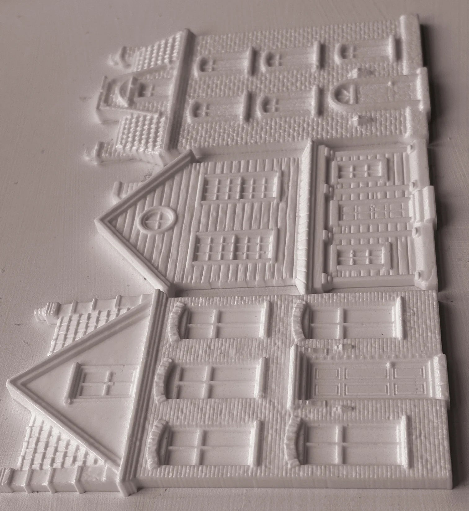 White plastic miniature building pieces for dioramas, featuring detailed house designs with windows, doors, and textured brick patterns, arranged on a light gray background.
