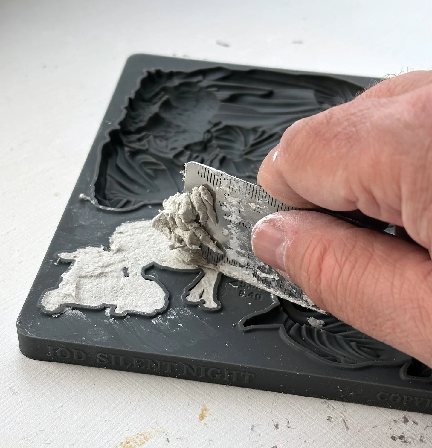 A close-up of a hand using a metal spatula to apply white silicone mold release agent onto a dark gray custom-shaped silicone mold, with the embossed text 'JOD SILENTNIGHT' visible on the bottom edge.
