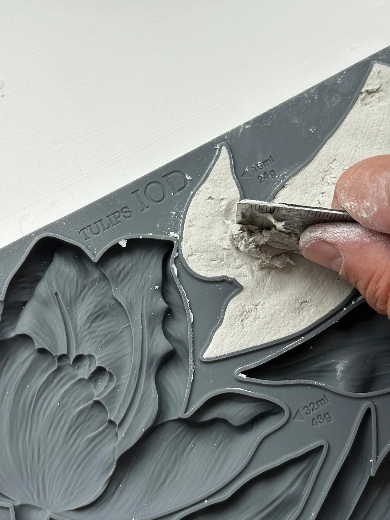 Hand using a tool to shape gray clay in a mold with leaf patterns.
