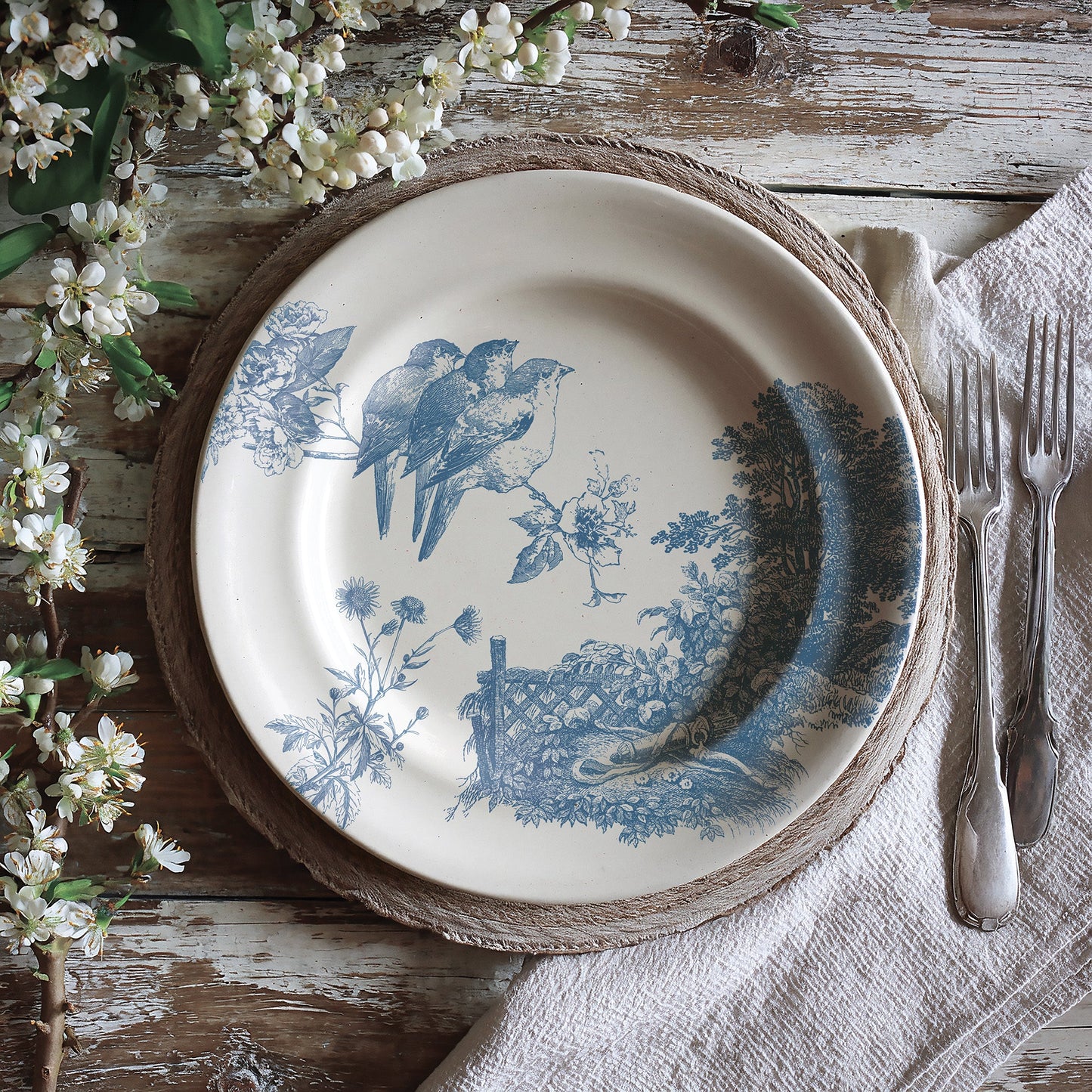 Decorative plate with blue bird design on a rustic wooden table with flowers and cutlery.
