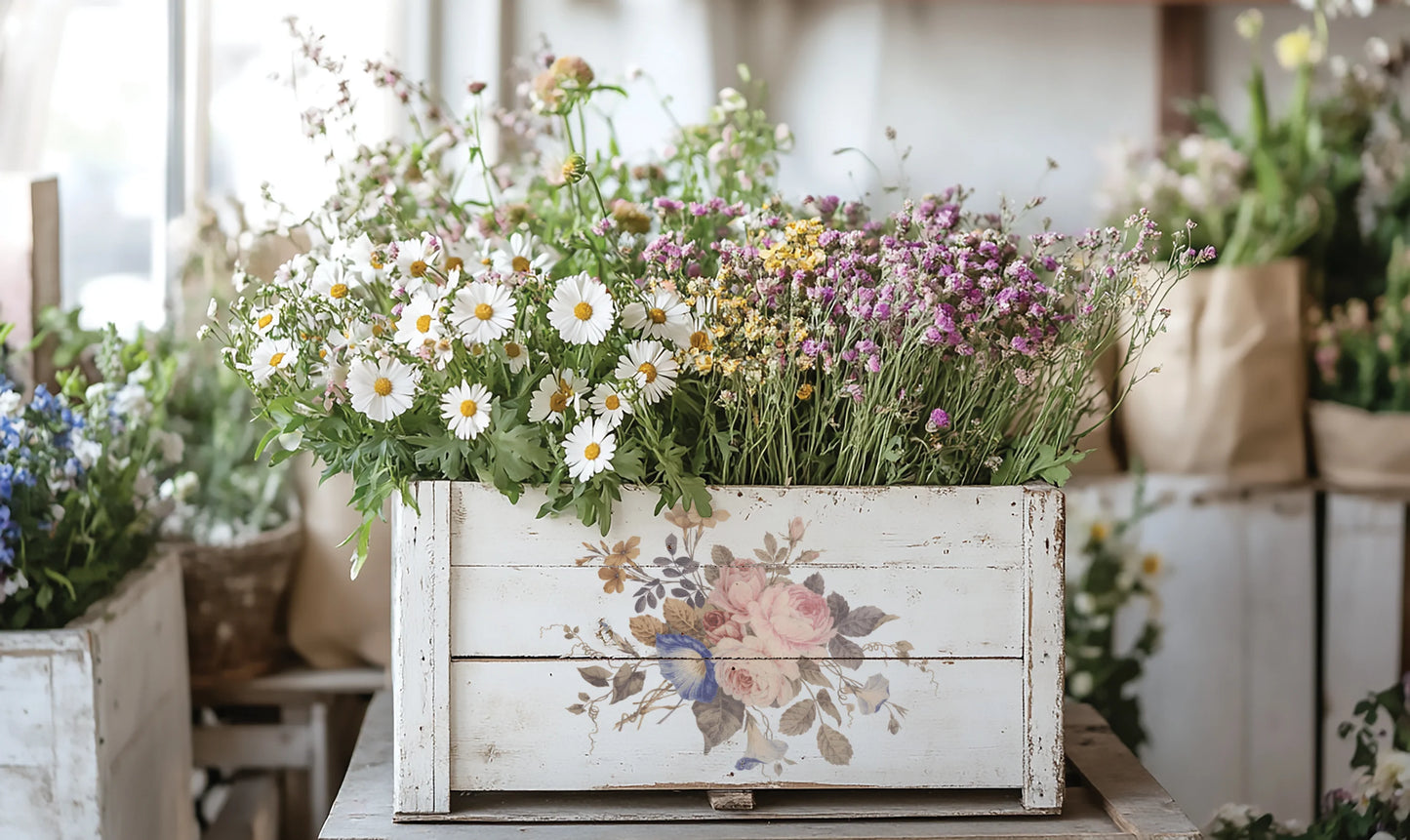 Floral arrangement in a decorative wooden box with a blurred background of more plants.