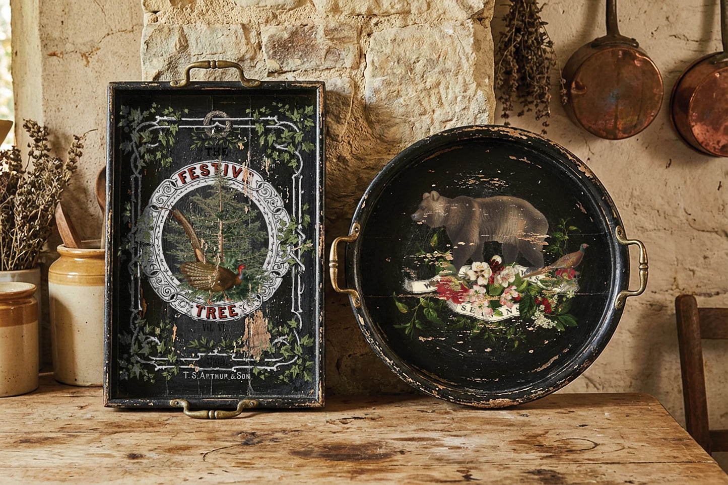 A rustic decorative serving tray set displayed on a wooden table against a stone wall. The set includes a rectangular black tray with ornate green vine borders and a central circular illustration, and a round black tray with brass handles, both featuring a painted bear and floral design.