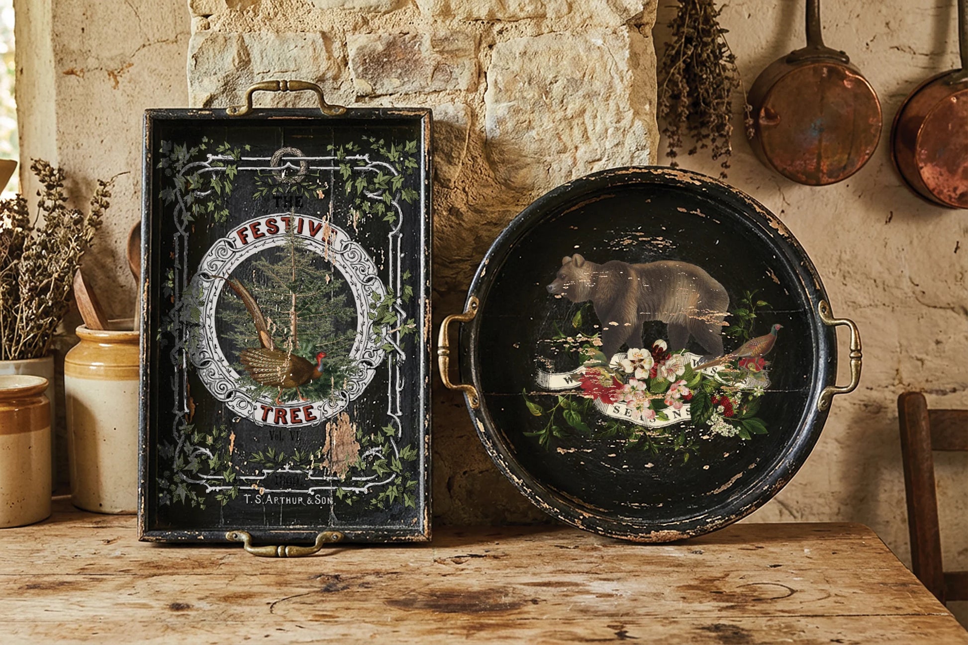 A rustic decorative serving tray set displayed on a wooden table against a stone wall. The set includes a rectangular black tray with ornate green vine borders and a central circular illustration, and a round black tray with brass handles, both featuring a painted bear and floral design.