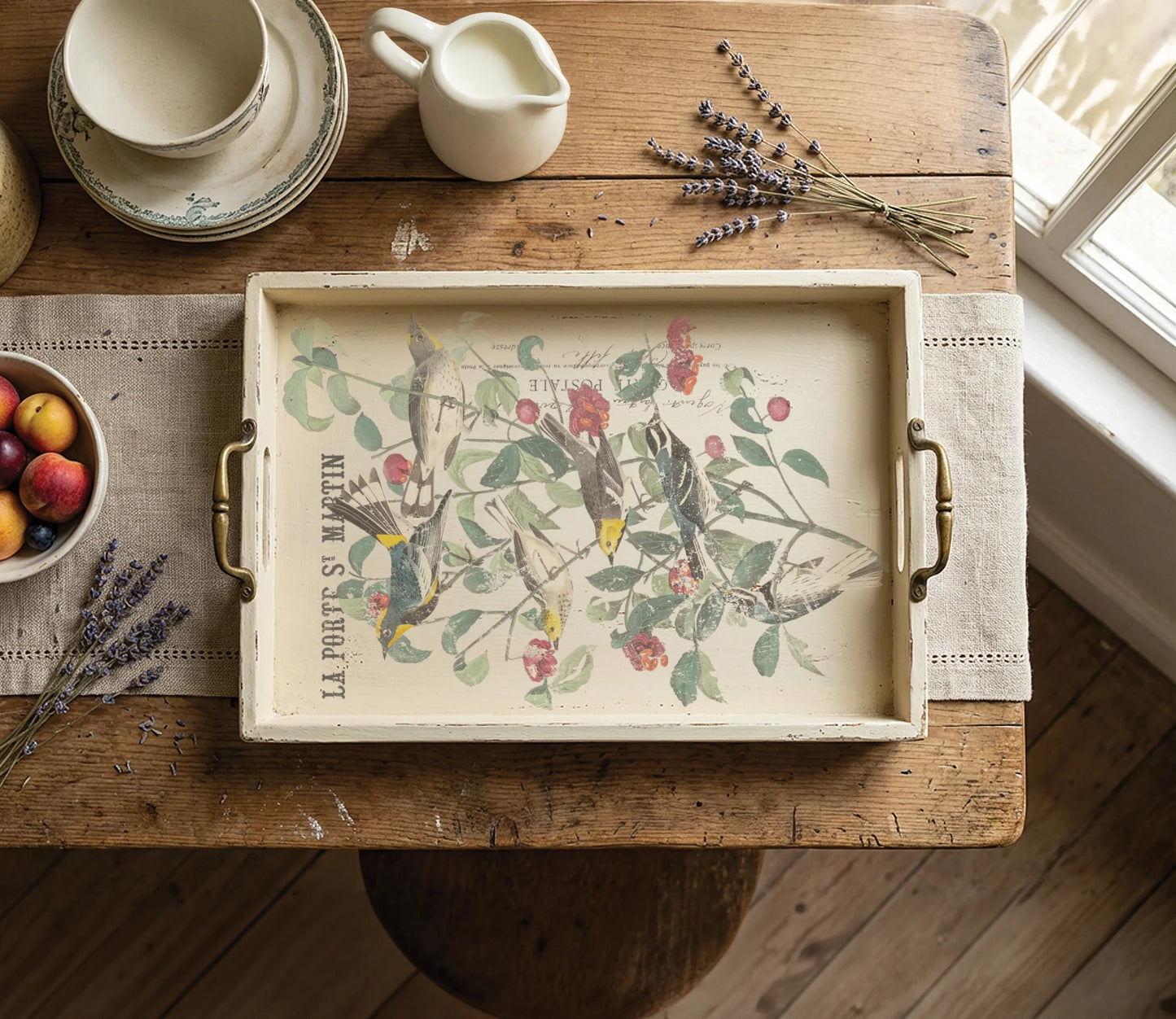 A rectangular serving tray with brass handles, featuring a vintage-style botanical illustration of birds and cherries with the text 'LA PORTS S. MARTIN', displayed on a rustic wooden table with a placemat, fruit bowl, lavender, and cream-colored ceramic items.