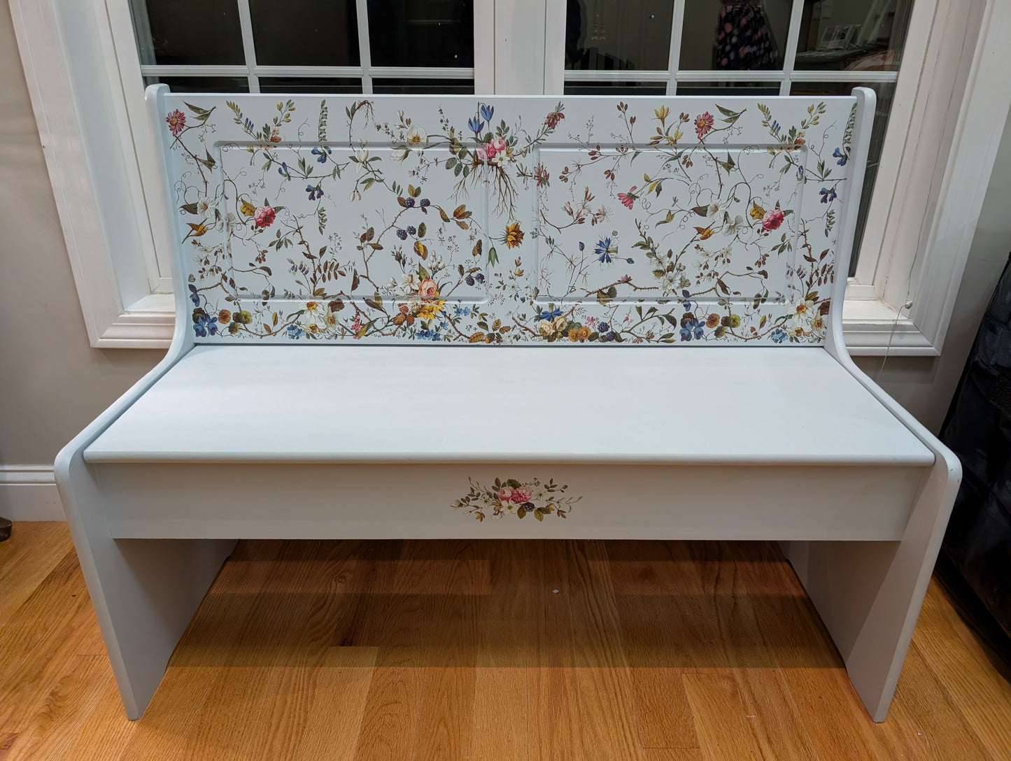 White bench with floral-patterned backrest in a room with wooden floor and window.