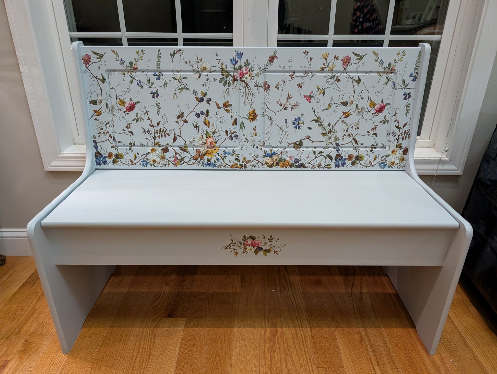 White bench with floral-patterned backrest in a room with wooden floor and window.