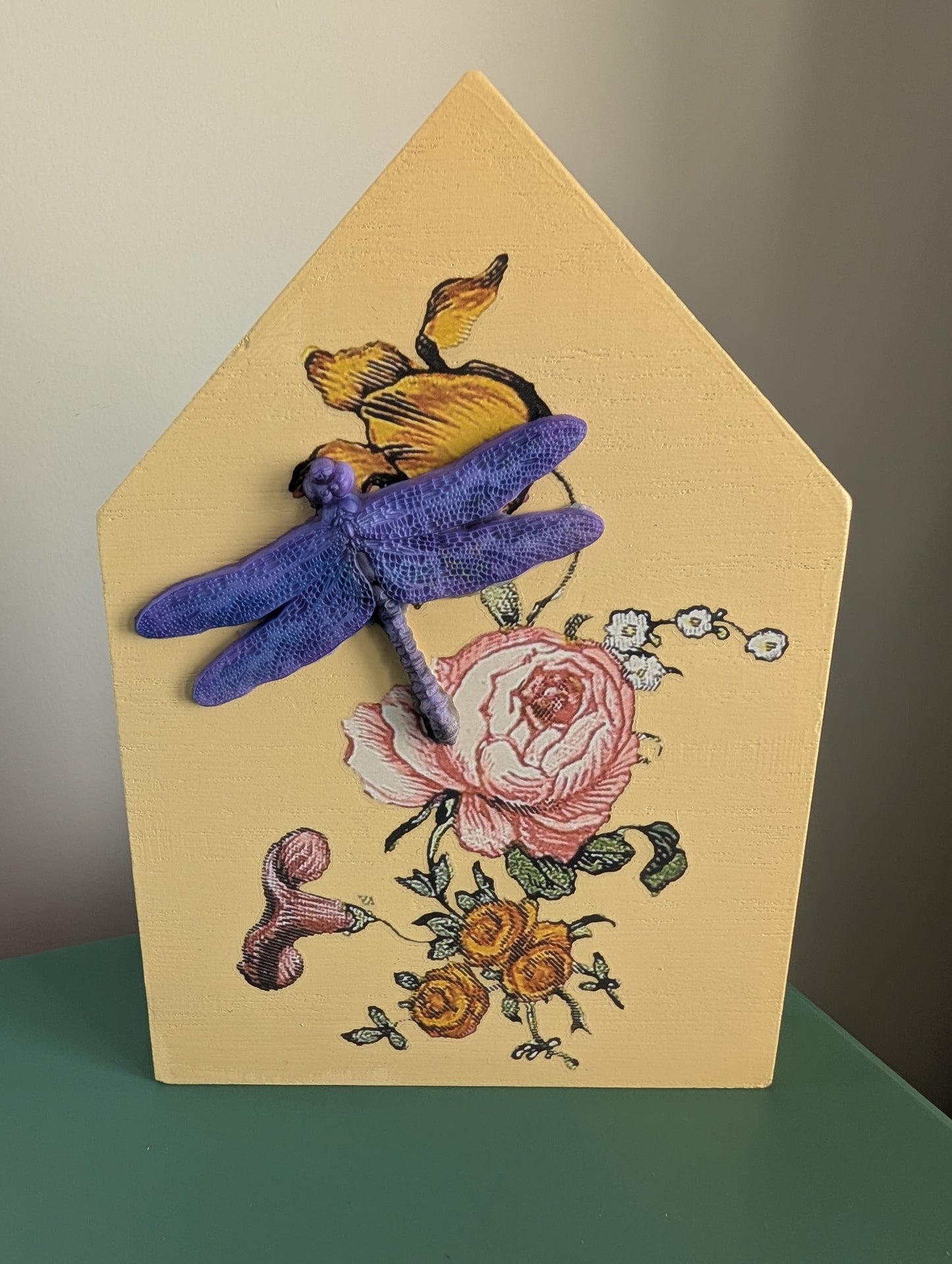Decorative house-shaped plaque with dragonfly and floral design on a beige background.